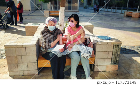 Mother and Daughter Rest After Flu Shots on Sunny Bench Outside Medical Center, Wearing Masks and Reflecting on Health and Safety Protocols. 117372149
