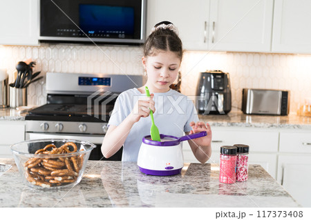 With focused attention, this budding culinary artist dips pretzels into a pot of melted chocolate, creating sweet delights in the warmth of a well-lit home kitchen. 117373408