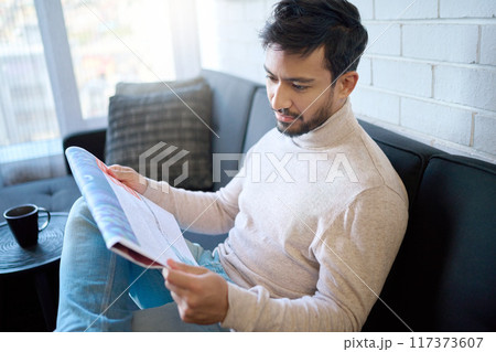 News, newspaper and man reading article in a home, house or apartment on the weekend sitting on a couch or sofa. Mexican, relax and young person enjoying free time in living room in the morning News, newspaper and man reading article in a home, house or apartment on the weekend sitting on a couch or sofa. Mexican, relax and young person enjoying free time in living room in the morning 117373607