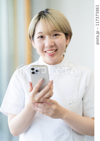 スマホを持つ金髪ショートヘアの若い女性整体師（髪色自由、自分らしく働く、職場環境） 117373861