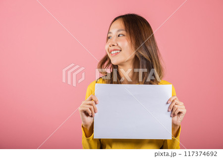 Portrait Asian happy beautiful young woman teen holding a blank sheet of white A4 paper poster for advertising studio shot isolated on pink background, female smiling with copy space 117374692