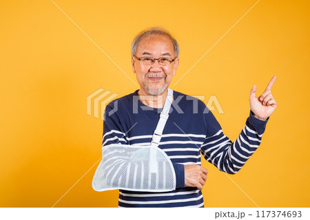 Portrait Asian old man cast on his arm is pointing space studio shot isolated yellow background, retired elderly man after accident broken arm wear arm splint for treatment accident insurance concept 117374693