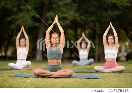 Portrait of women sitting on gym mat and doing yoga meditation in lotus position and arms raised in park Portrait of women sitting on gym mat and doing yoga meditation in lotus position and arms raised in park 117376158