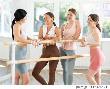 Young ballerinas chatting joyfully standing around training barre 117376471