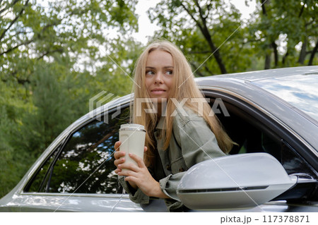 Blonde woman sticking head out of windshield car and drink coffee or tea from reusable mug. Young tourist explore local travel making candid real moments. Refuse reuse recycle zero waste concept 117378871