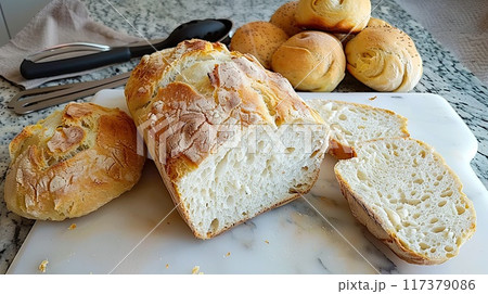 A loaf of freshly baked artisan bread sliced open, showcasing its airy texture, placed on a cutting board with rolls in the background.. 117379086