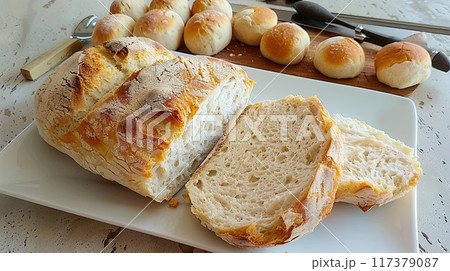 A loaf of freshly baked artisan bread sliced open, showcasing its airy texture, placed on a cutting board with rolls in the background.. A loaf of freshly baked artisan bread sliced open, showcasing its airy texture, placed on a cutting board with rolls in the background.. 117379087