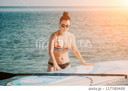 Sea woman sup. Silhouette of happy middle aged tanned woman in rainbow bikini, surfing on SUP board, confident paddling through water surface. Idyllic sunset. Active lifestyle at sea or river. Sea woman sup. Silhouette of happy middle aged tanned woman in rainbow bikini, surfing on SUP board, confident paddling through water surface. Idyllic sunset. Active lifestyle at sea or river. 117380489