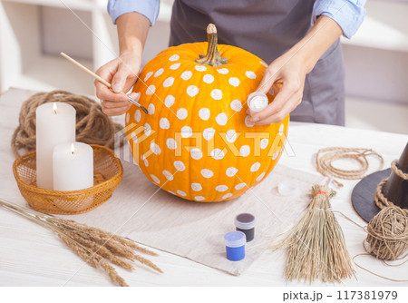 Woman in apron painting Halloween pumpkin, at wooden table, preparing holiday decorations at home 117381979