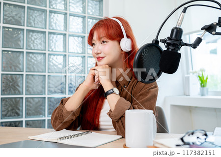 A beautiful Asian podcaster is in her studio, looking away and smiling with a dreamy expression. 117382151
