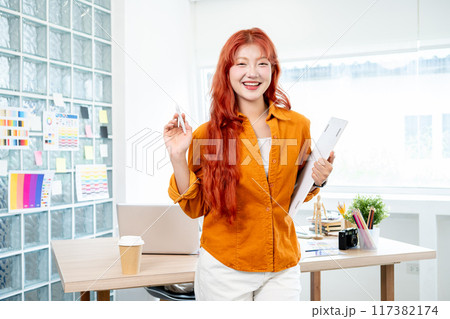 An attractive Asian female graphic designer is standing in her studio and smiling at the camera. 117382174