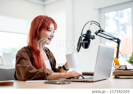 A beautiful Asian female podcaster is working in her home studio and enjoying a coffee. 117382205