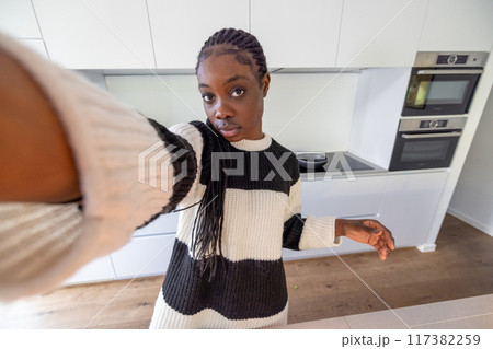 A young woman in a modern kitchen, wearing a striped sweater, captures a selfie 117382259
