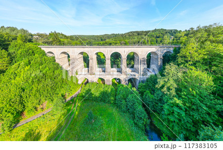The Sychrov Railway Bridge stands majestically with graceful arches, surrounded by lush greenery. The bright sky enhances the tranquil setting. 117383105