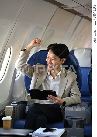 Young businessman sitting in airplane seat and celebrating success with his arm raised in triumphant gesture 117384195