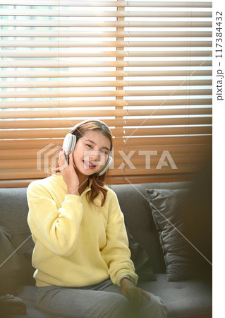 Happy young Asian woman enjoying music over headphones while relaxing on sofa at home Happy young Asian woman enjoying music over headphones while relaxing on sofa at home 117384432