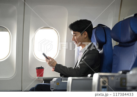 Side view of asian businessman sitting on the aircraft seat near the window and using mobile phone Side view of asian businessman sitting on the aircraft seat near the window and using mobile phone 117384433