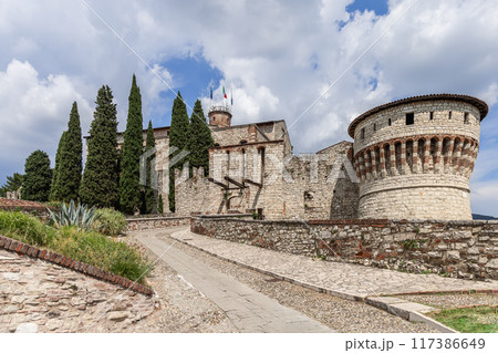 Brescia medieval castle entrance with drawbridge, stone pathway, and cypress trees under cloudy sky Brescia medieval castle entrance with drawbridge, stone pathway, and cypress trees under cloudy sky 117386649