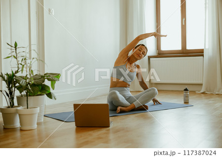 A woman is diligently practicing her yoga routine at home while actively engaging with her laptop A woman is diligently practicing her yoga routine at home while actively engaging with her laptop 117387024