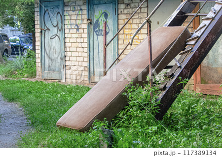 Old brown textured mattress leaning on metal stairs left outside near brick wall shed with wooden doors and green grass around Old brown textured mattress leaning on metal stairs left outside near brick wall shed with wooden doors and green grass around 117389134