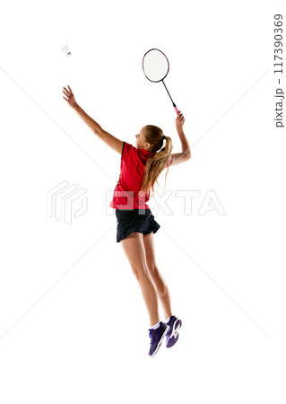 Side view photo of focused on game young woman, badminton player serving shuttlecock against white studio background. Side view photo of focused on game young woman, badminton player serving shuttlecock against white studio background. 117390369
