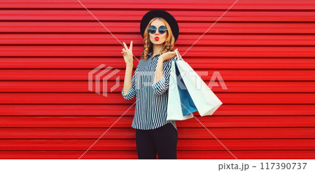 Portrait beautiful young woman with shopping bags blowing kiss in shirt, black hat on red background Portrait beautiful young woman with shopping bags blowing kiss in shirt, black hat on red background 117390737