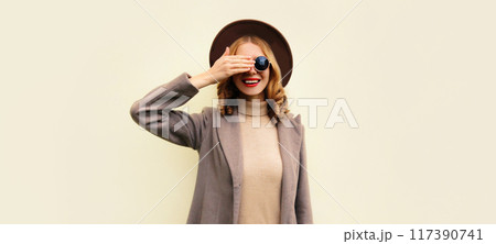Portrait of beautiful smiling young woman model wearing brown round hat and coat on beige background 117390741