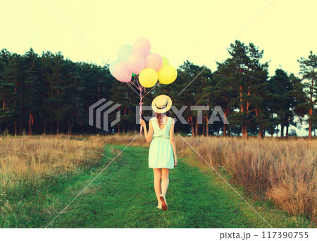 Rear view of young woman with bunch of colorful balloons in summer hat, dress in field outdoors 117390755