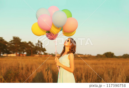 Happy cute smiling young woman with bunch of colorful balloons wearing dress outdoors 117390758