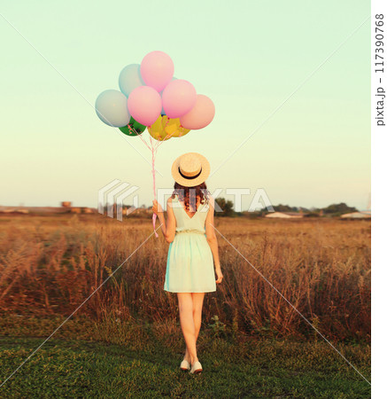 Rear view of young woman with bunch of colorful balloons in summer hat, dress in field outdoors 117390768