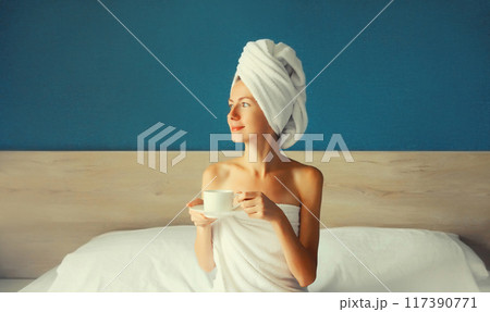 Happy young woman enjoying with cup of coffee sitting on bed looking out the window in the morning Happy young woman enjoying with cup of coffee sitting on bed looking out the window in the morning 117390771