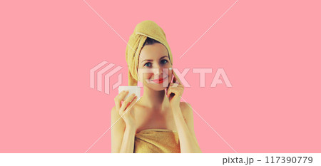 Portrait happy smiling young woman applying face cream drying hair in bath towel on pink background Portrait happy smiling young woman applying face cream drying hair in bath towel on pink background 117390779