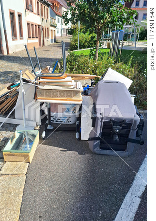 Discarded items including overturned office chair, wooden boards, home furnishings cluttering sidewalk, reflecting urban waste management issues, necessity for responsible disposal. Vertical picture. Discarded items including overturned office chair, wooden boards, home furnishings cluttering sidewalk, reflecting urban waste management issues, necessity for responsible disposal. Vertical picture. 117391349