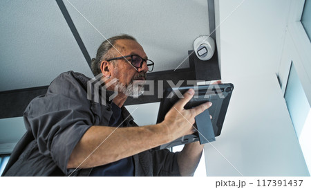 Male mature installer sets up CCTV camera in office room using tablet computer Male mature installer sets up CCTV camera in office room using tablet computer 117391437