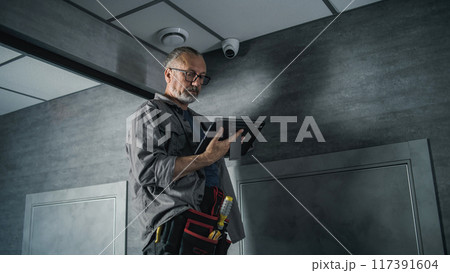 Male installer adjusts the angle of CCTV camera in office, uses tablet computer 117391604