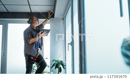 Male technician stands on ladder, installs CCTV camera in office room using tablet Male technician stands on ladder, installs CCTV camera in office room using tablet 117391670