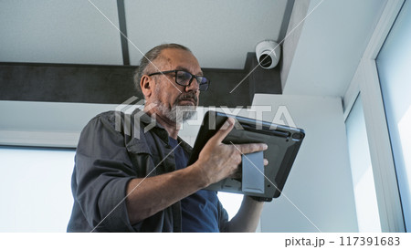Male mature installer sets up CCTV camera in office room using tablet computer 117391683