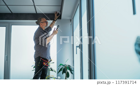 Male mature installer adjusts security camera in office using mobile phone 117391714