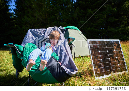 Young child plays with smartphone while charging with photovoltaic solar panel near tourist tent in summer. Integration of renewable energy in outdoor camping activities. 117391758