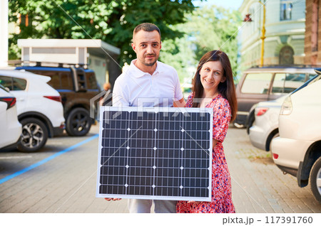 Cute couple holding portable solar panel. Young man and his wife showing their green lifestyle. Concept of alternative and renewable energy sources. Cute couple holding portable solar panel. Young man and his wife showing their green lifestyle. Concept of alternative and renewable energy sources. 117391760