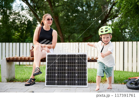Woman sits on park bench beside solar panel while young boy wearing helmet runs joyfully nearby. Integration of renewable energy into everyday life, emphasizing sustainability environments. 117391761