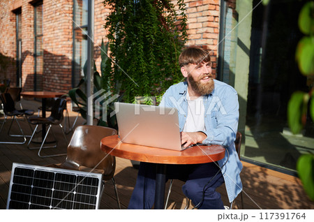 Bearded happy man works on laptop at outdoor round table, with glass of iced coffee. Solar panel nearby, emphasizing sustainable, modern eco-friendly workspace combines technology and sustainability. 117391764