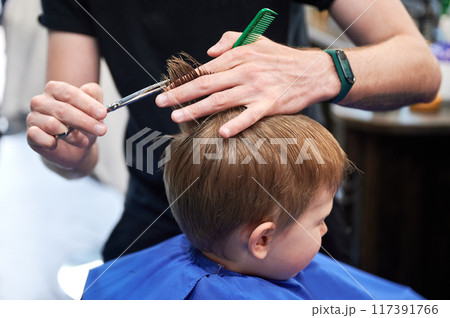 Man barber cutting little boy's hair using comb and scissors. Child getting haircut from adult male, likely barber. Professional hairdresser and cute client at modern barbershop. Man barber cutting little boy's hair using comb and scissors. Child getting haircut from adult male, likely barber. Professional hairdresser and cute client at modern barbershop. 117391766
