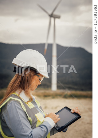 A skilled female engineer uses a modern tablet at a vast wind farm, highlighting the blend of advanced technology and innovative engineering in a sustainable, ecofriendly environment 117391963
