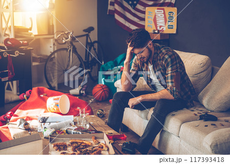Frustrated young man keeping hand in hair while sitting on sofa in messy room after party 117393418