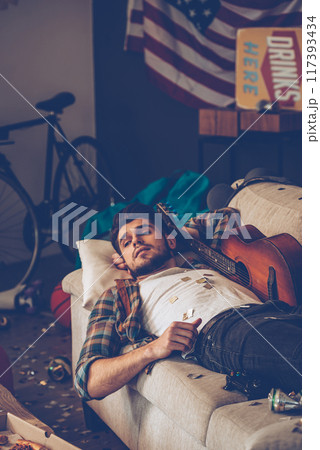 Young handsome man looking away while lying down on sofa in messy room after party Young handsome man looking away while lying down on sofa in messy room after party 117393434