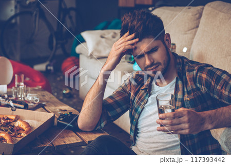 Handsome young man holding glass of water and looking exhausted while sitting on the floor in messy room after party Handsome young man holding glass of water and looking exhausted while sitting on the floor in messy room after party 117393442