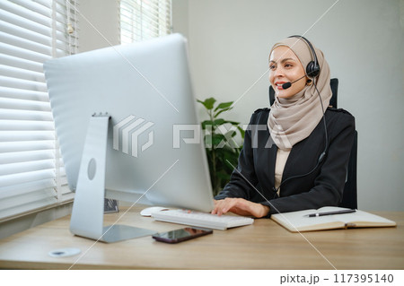 A woman wearing a headset is sitting at a desk in front of a computer monitor A woman wearing a headset is sitting at a desk in front of a computer monitor 117395140