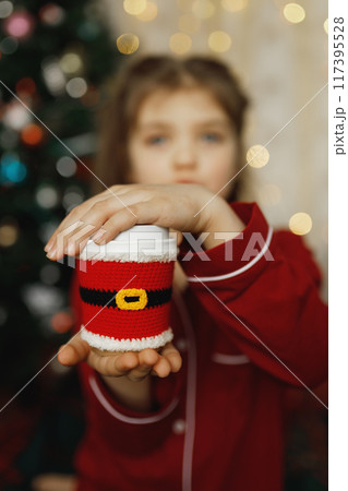 cup of coffee in knitted clothes of Santa Claus in hands of child in red pajamas 117395528