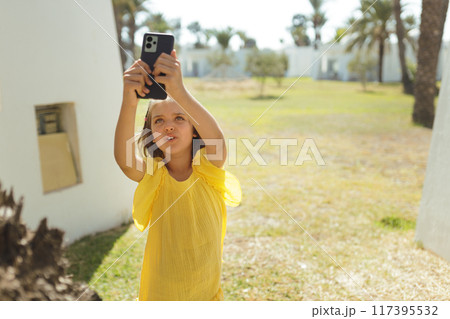 Girl 7 years old in yellow dress takes photos on phone on lawn with palm trees in summer day 117395532